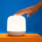 A small cylindrical smart lamp with a glowing white shade and a white base sits on an orange platform against a blue background, with a human hand reaching toward the top.