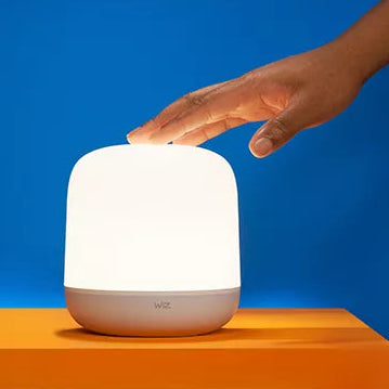 A small cylindrical smart lamp with a glowing white shade and a white base sits on an orange platform against a blue background, with a human hand reaching toward the top.