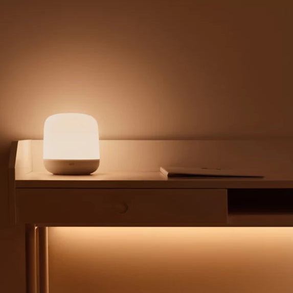 A small cylindrical table lamp with a frosted white top and base, glowing warm light, sitting on a minimalist wooden desk against a warm brown wall.