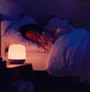 A small glowing bedside lamp on a nightstand next to a bed where a person is sleeping under white bedding in a dimly lit bedroom.